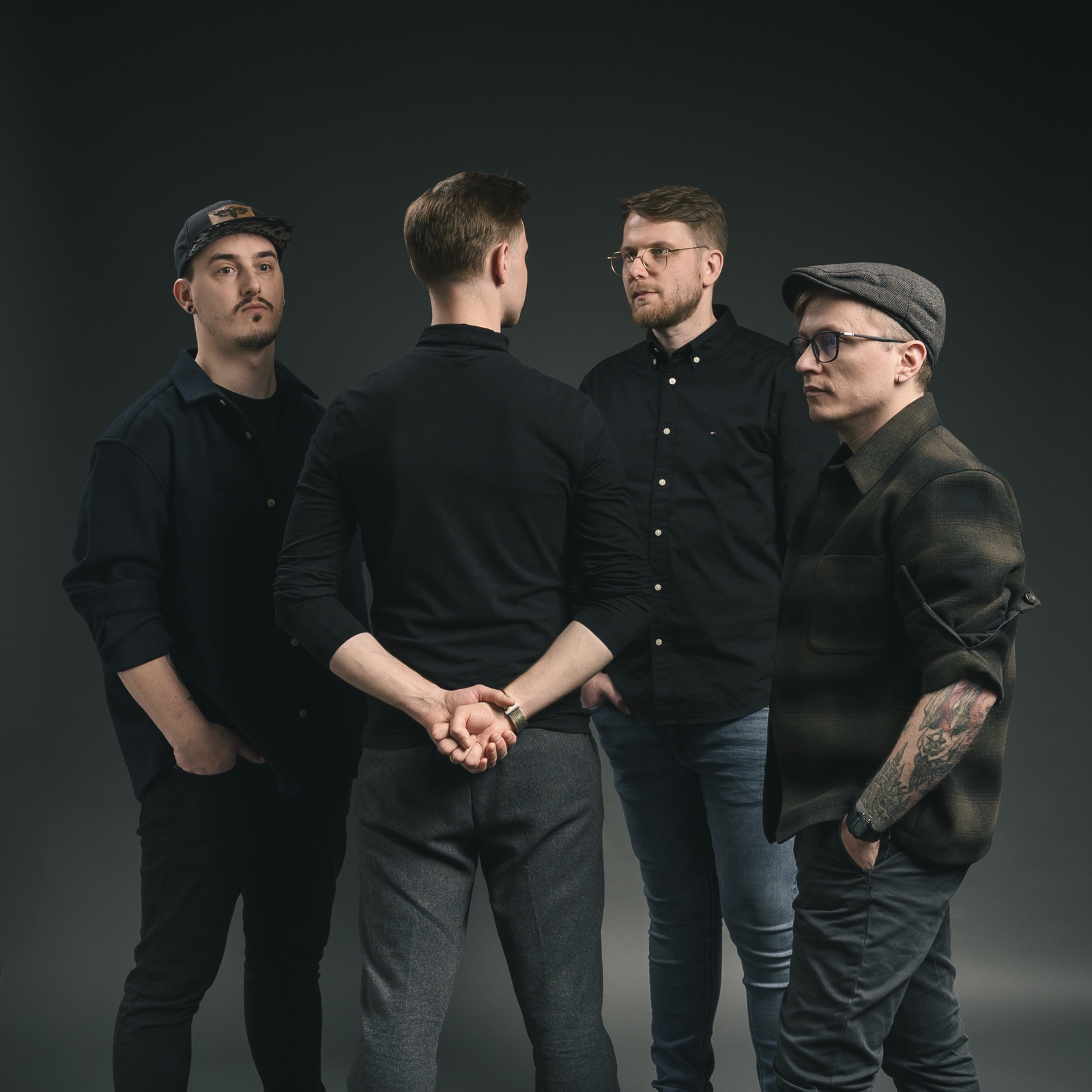 Gruppenbild im Studio vor dunkelgrauem Hintergrund: Ein Bandmitglied steht mit dem Rücken zur Kamera, die drei anderen stehen ihm zugewandt.