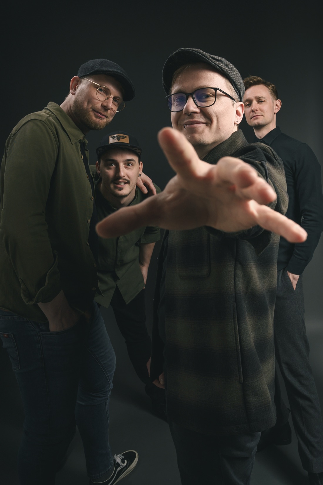 Ein Bandmitglied streckt die Hand in die Kamera, die drei anderen dahinter im Studiolicht.